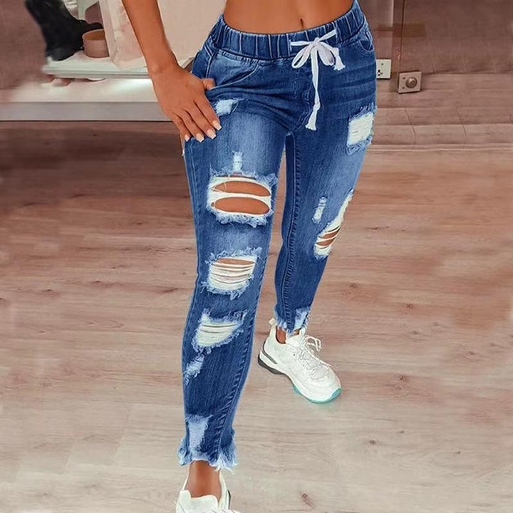 Close-up of a woman wearing ripped blue denim jogger jeans with white drawstring and white sneakers on wooden floor