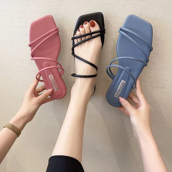 Three strappy square-toe flat sandals in black on foot, pink and blue held by hands on beige background
