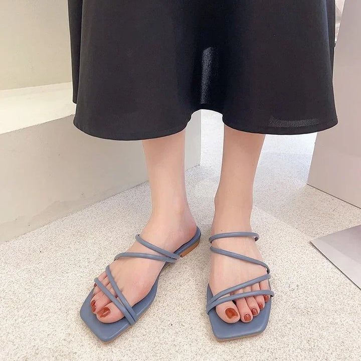 Feet wearing stylish blue strappy square-toe sandals with red nail polish on beige floor