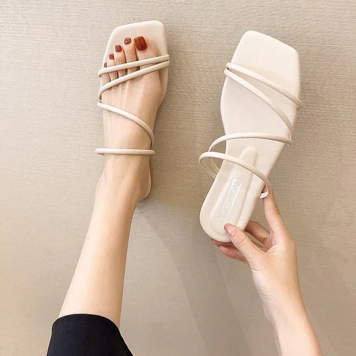 Woman wearing one beige strappy square-toe sandal, holding the matching sandal against beige background