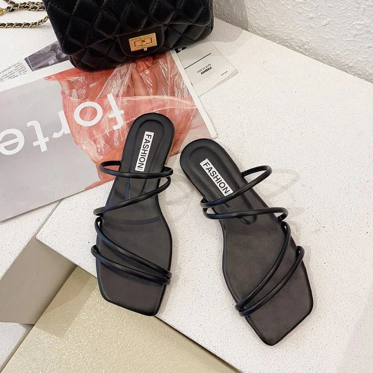 black strappy flat sandals with square toe on a white surface next to a quilted black handbag and fashion magazine
