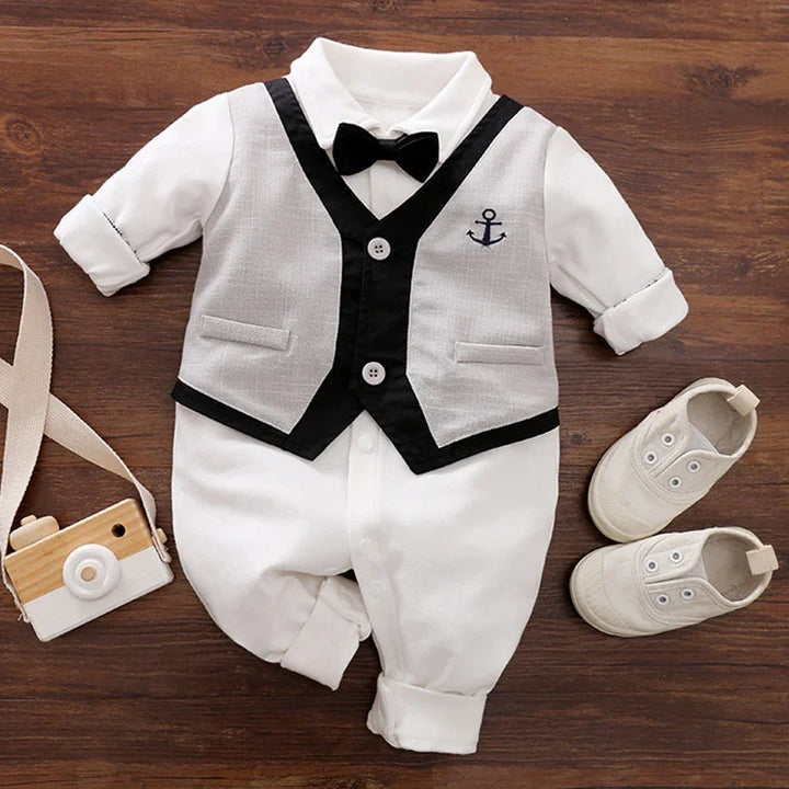 Elegant baby boy outfit with white jumpsuit, gray vest with anchor emblem, black bow tie, and white shoes on wooden background
