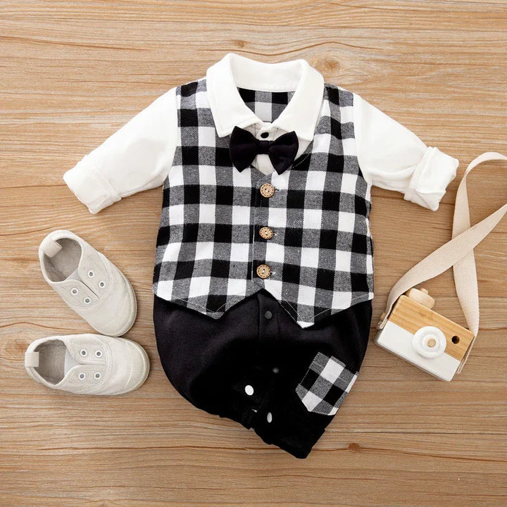 Black and white checkered baby romper with bow tie, white shirt, beige baby shoes, wooden toy camera on wooden background