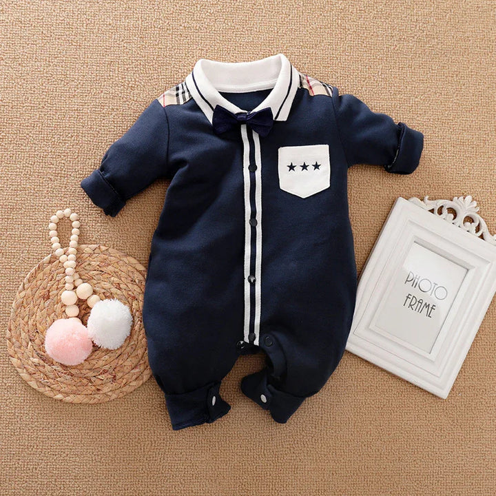 Navy blue baby romper with white collar, bow tie, star patch, and striped button placket on beige carpet