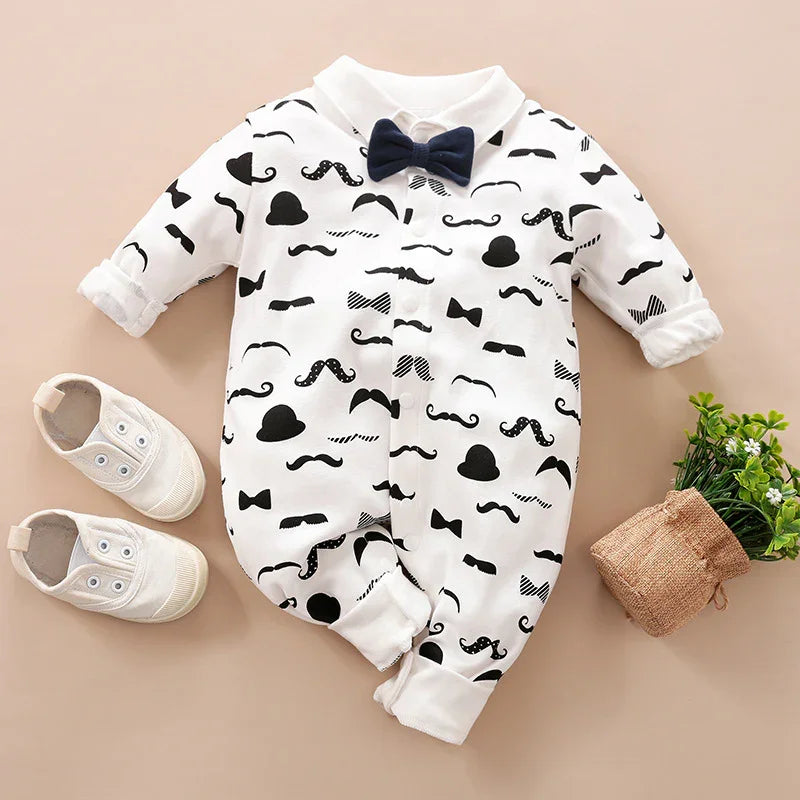 White baby romper with black hat and mustache prints, navy blue bow tie, paired with white baby shoes and small potted plant