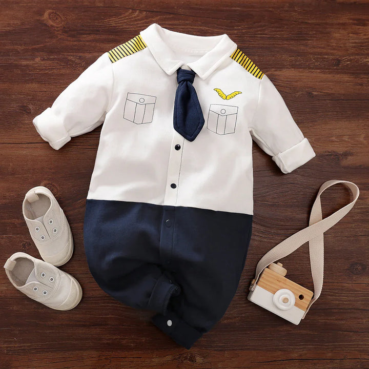 Baby pilot costume romper with navy tie and yellow-striped epaulets, paired with beige shoes on wooden floor