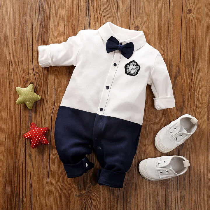 Baby boy formal romper with white top and navy pants, navy bow tie, white shoes on wooden floor