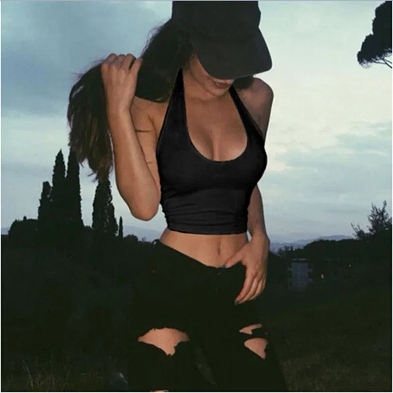Woman in black sports crop top and ripped black jeans outdoors at dusk with cap and long hair