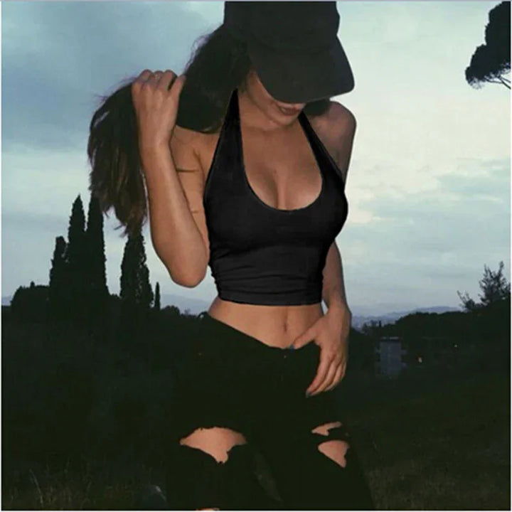 Woman in black sports crop top and ripped black jeans outdoors at dusk with cap and long hair
