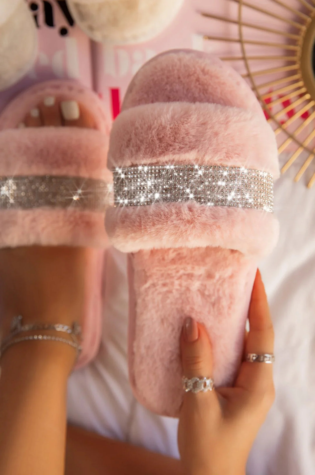 Soft pink fuzzy slippers with sparkling rhinestone straps, worn on feet with painted nails and silver rings