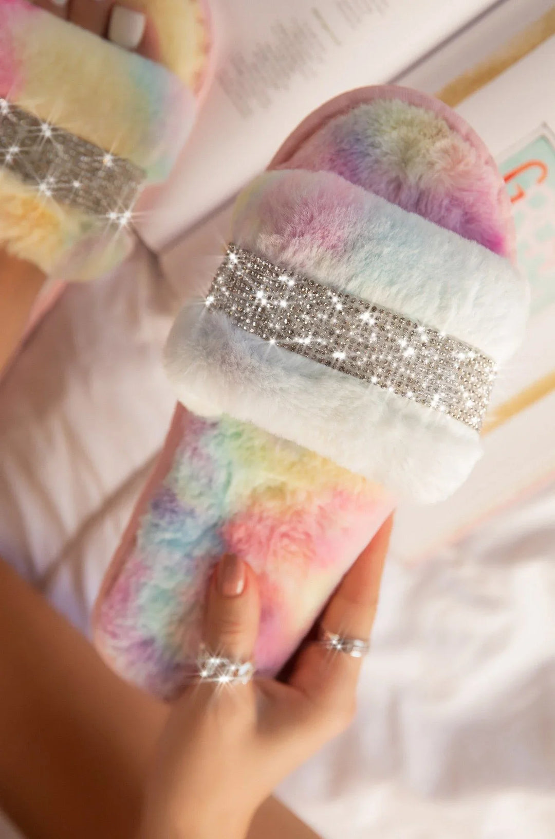 Hand holding rainbow tie-dye fuzzy slippers with sparkling rhinestone strap on white bed