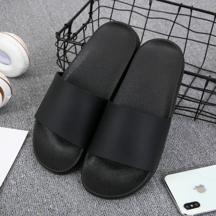 Pair of black slide sandals on gray carpet near a black wire basket and white smartphone