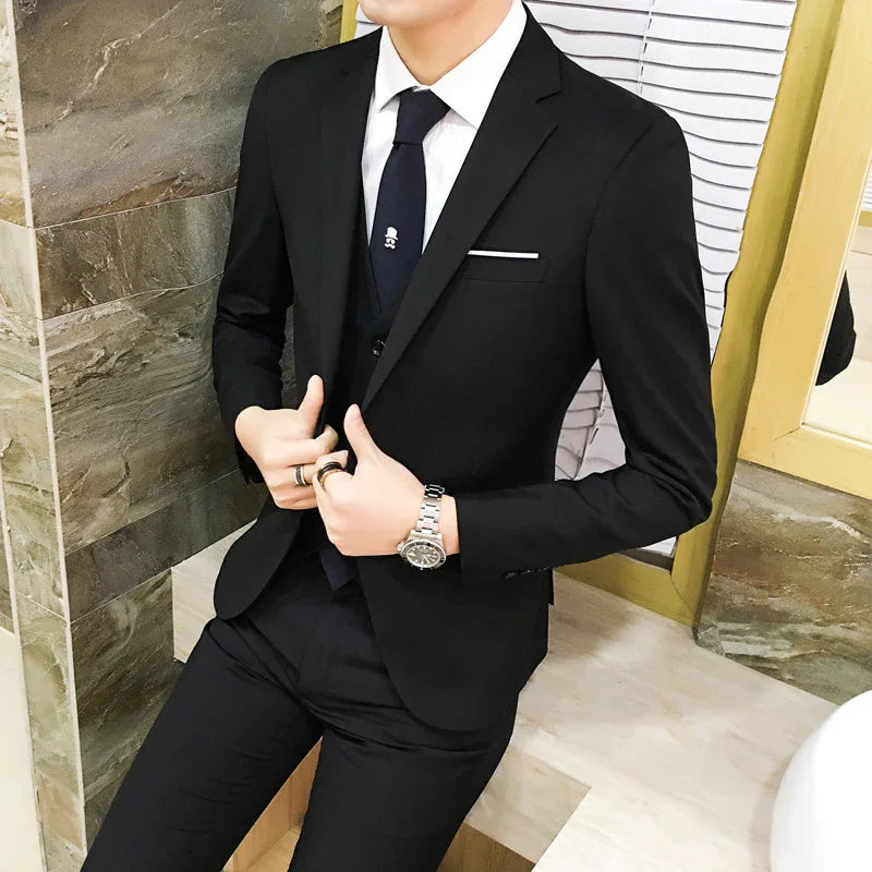 Man wearing a black slim-fit suit with white shirt and black tie sitting indoors by a marble wall
