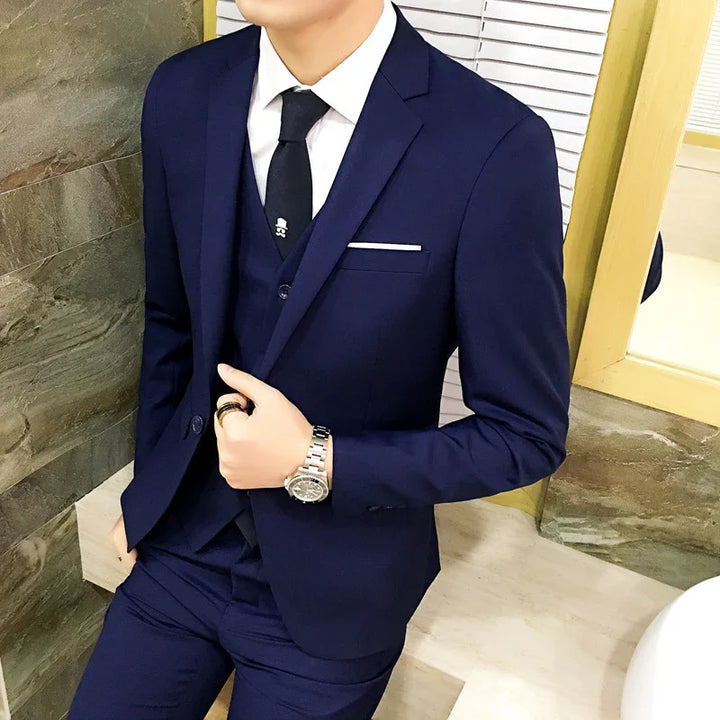 Man wearing a navy blue suit with white shirt, black tie, and silver wristwatch indoors