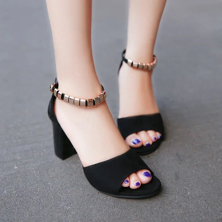 Black high heel sandals with gold square ankle straps and purple toenail polish on asphalt