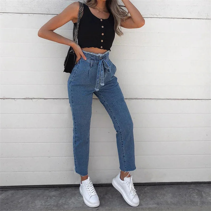 Woman wearing high-waisted blue jeans with tie belt, black buttoned crop top, white sneakers, and black shoulder bag against white wall