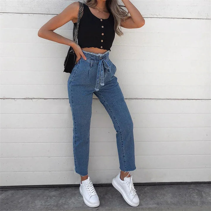 Woman wearing high-waisted blue jeans with tie belt, black buttoned crop top, white sneakers, and black shoulder bag against white wall
