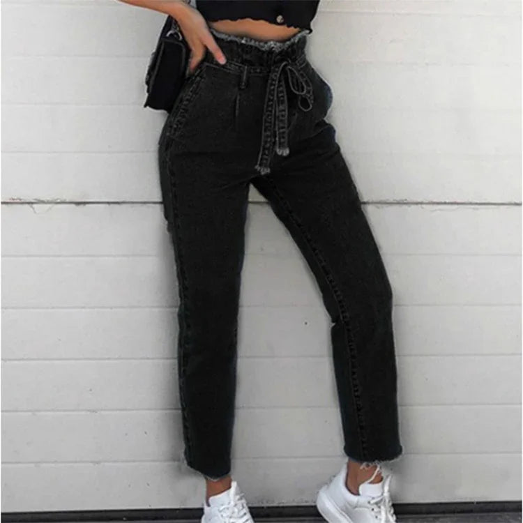Woman wearing high-waisted black denim jeans with paper bag waist and white sneakers against white brick wall