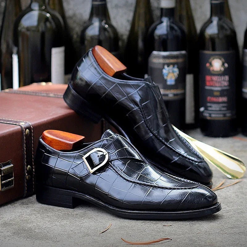 Black crocodile leather men's monk strap dress shoes with wooden shoe trees, vintage wine bottles and leather suitcase background