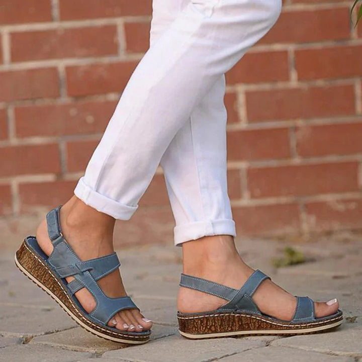 Person wearing blue wedge sandals with cork soles and white rolled-up pants on pavement