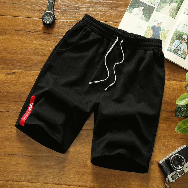 Black casual shorts with elastic waist and white drawstrings on wooden surface with watch, camera, magazine, and plant