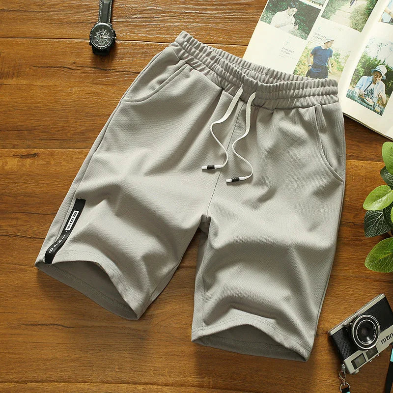 Light gray casual men's shorts with drawstring waistband on wooden surface with watch, camera, and magazine