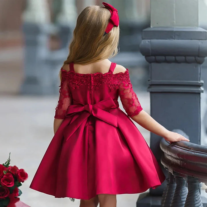 Little girl wearing elegant red lace off-shoulder dress with bow, standing outdoors near railing