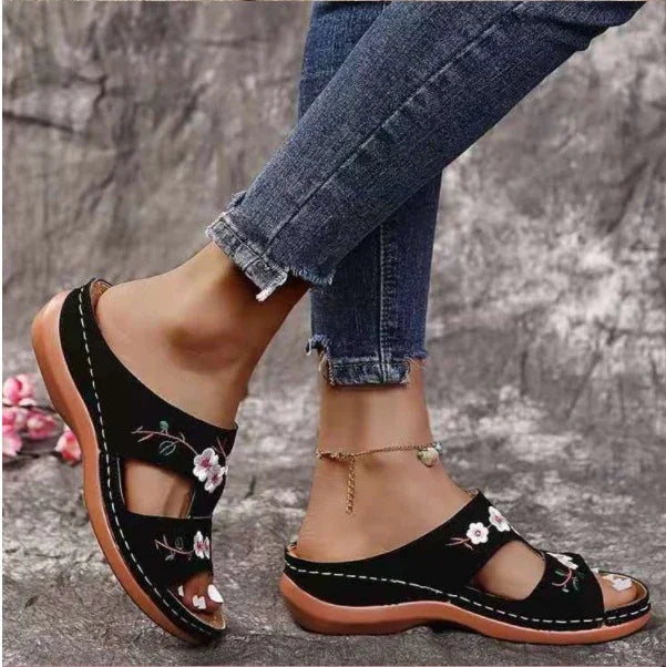 Black slip-on sandals with floral embroidery worn with ankle bracelet and frayed hem jeans on stone surface