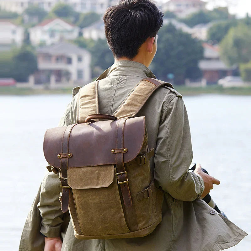 Man wearing khaki jacket and vintage canvas leather backpack by a lake with blurred houses in background