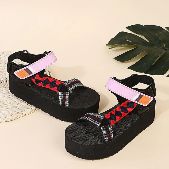 Black platform sandals with red patterned straps and pink ankle straps on beige background with green leaf