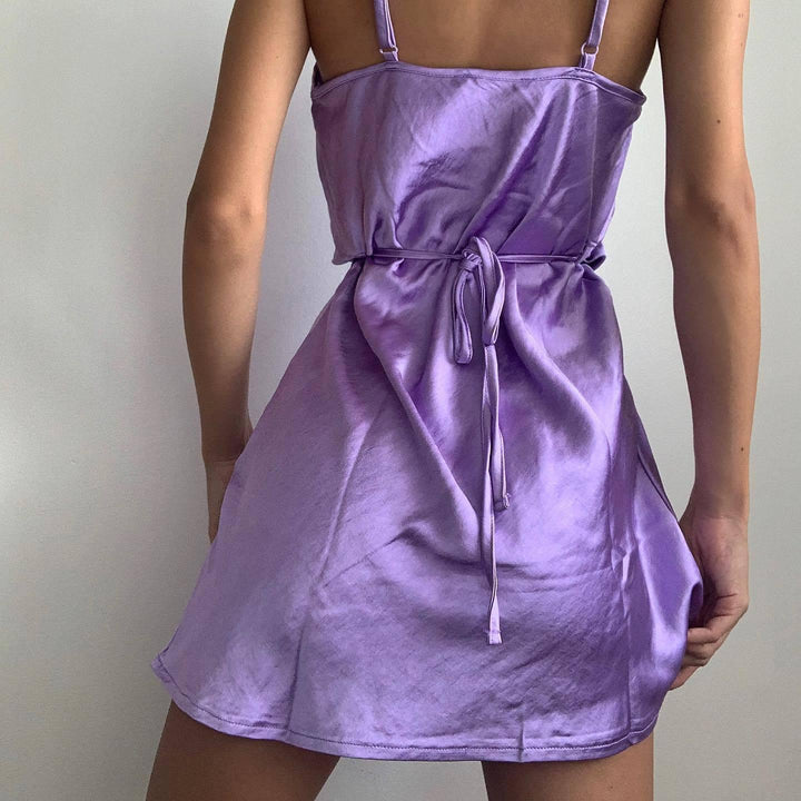 Back view of a woman wearing a short purple satin slip dress with thin straps and a tied waist belt