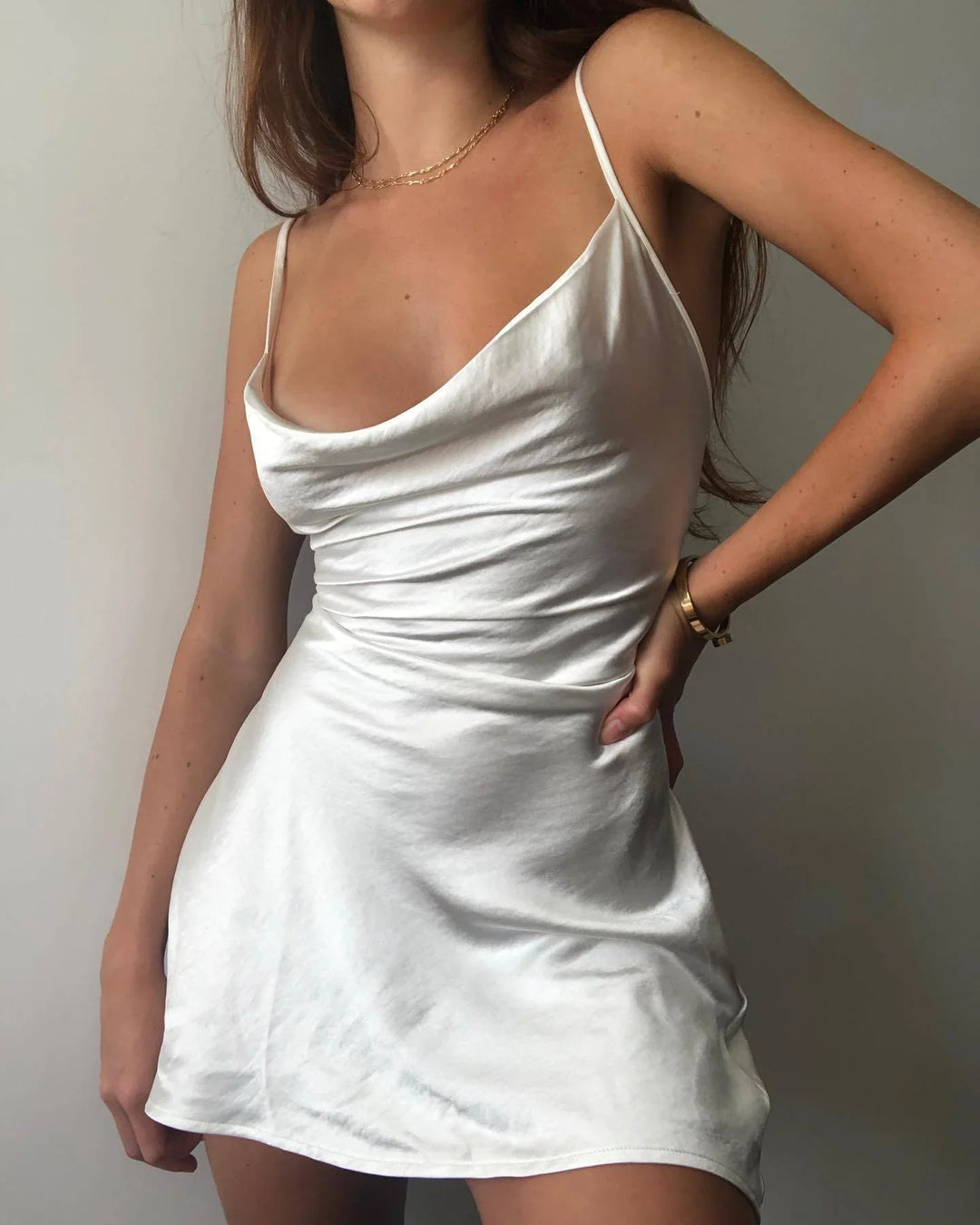 Woman wearing a white satin slip dress with thin straps and a gold bracelet against a plain background