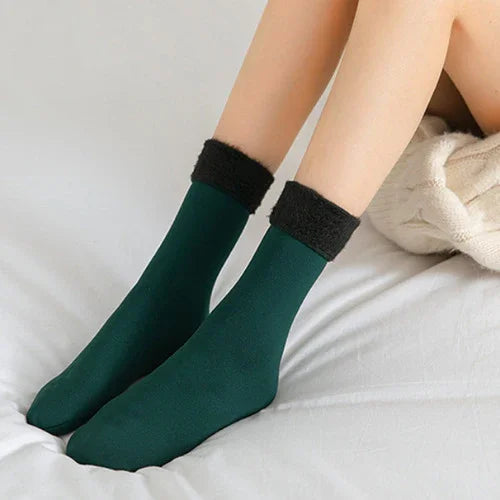 Close-up of feet wearing dark green warm socks with black fleece cuffs on a white bed