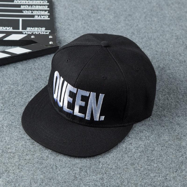Black baseball cap with bold white QUEEN. embroidery on front, placed on gray fabric surface