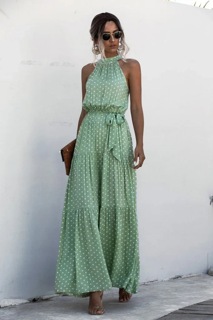Woman wearing sleeveless green polka dot maxi dress with tie waist, sunglasses, and holding brown clutch