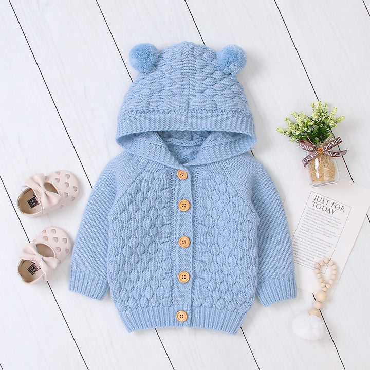 Blue knitted baby cardigan with hood and pom-poms, wooden buttons, surrounded by baby shoes and decorative flowers