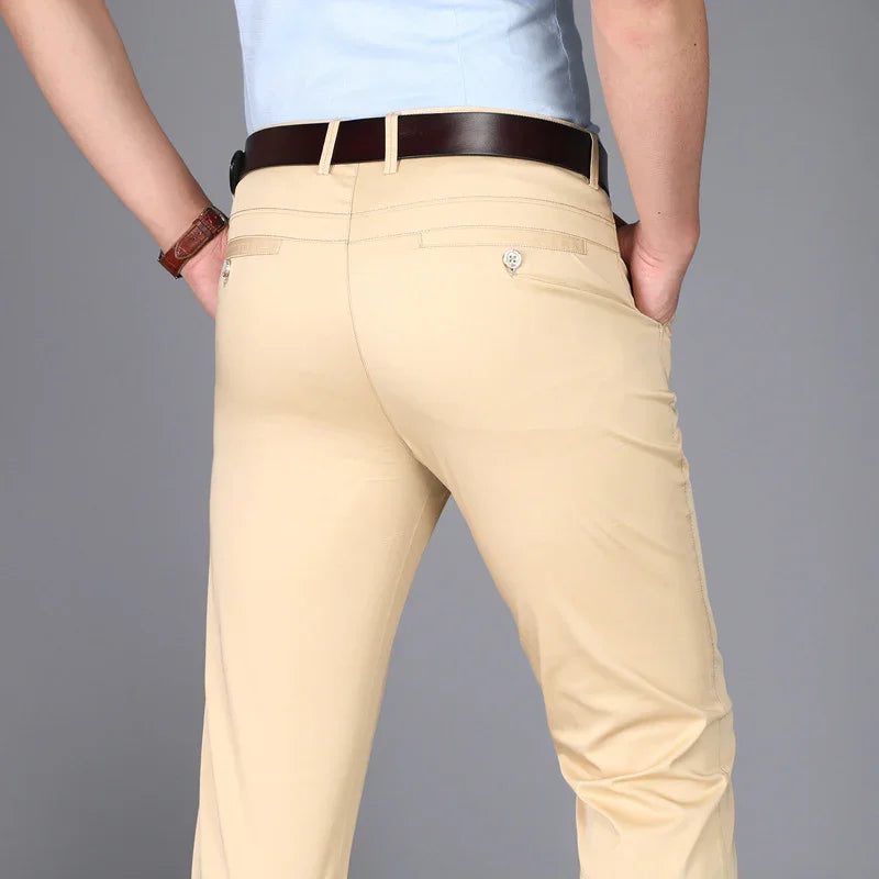 Man wearing beige chino pants with belt, hands in pockets, and light blue shirt against gray background