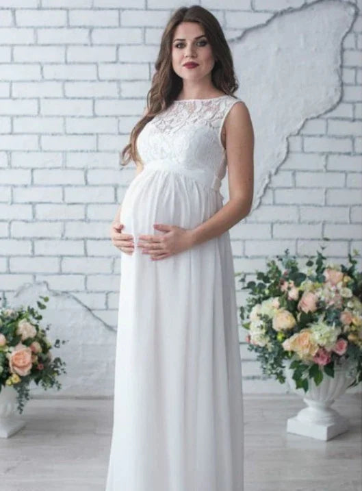 Pregnant woman in white lace gown posing indoors with floral arrangements and white brick wall backdrop