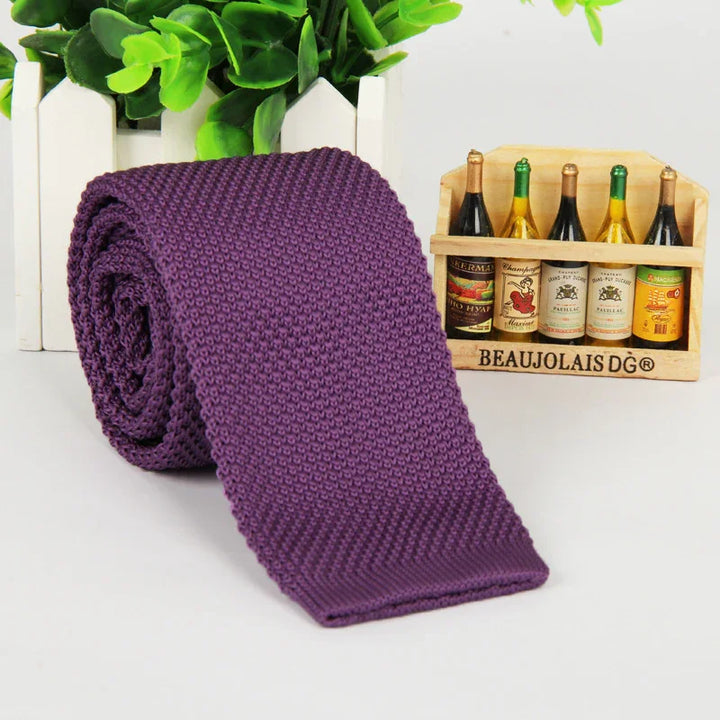 Rolled purple knitted tie placed on white surface with small wooden crate of miniature wine bottles and green plant background