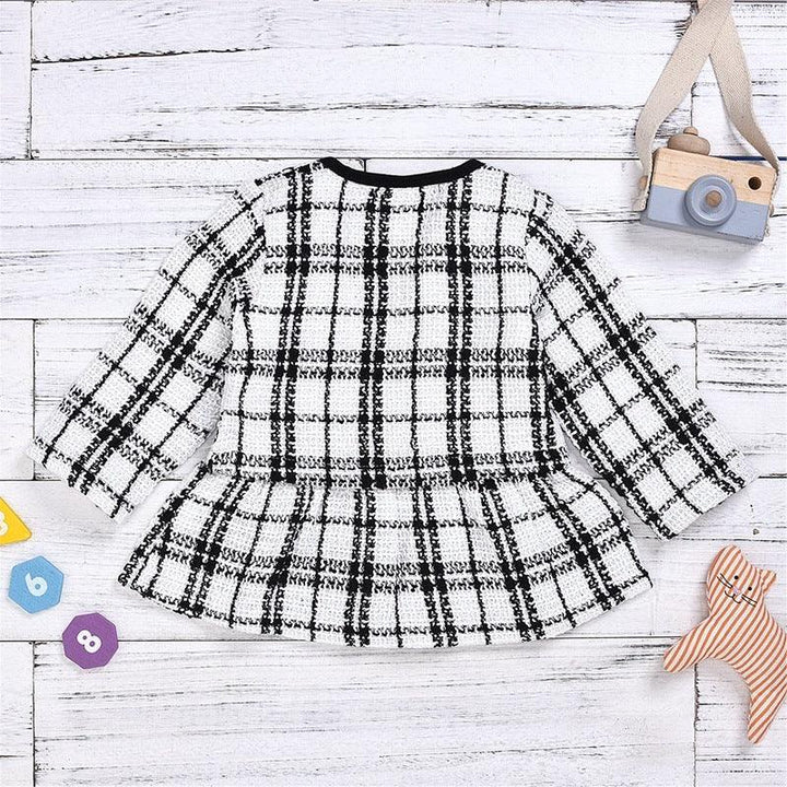 Black and white plaid long-sleeve toddler dress on white wooden background with toy camera and plush cat