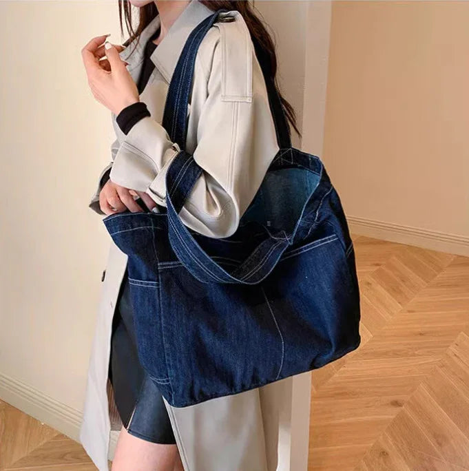 woman wearing beige trench coat holding large dark blue denim tote bag indoors