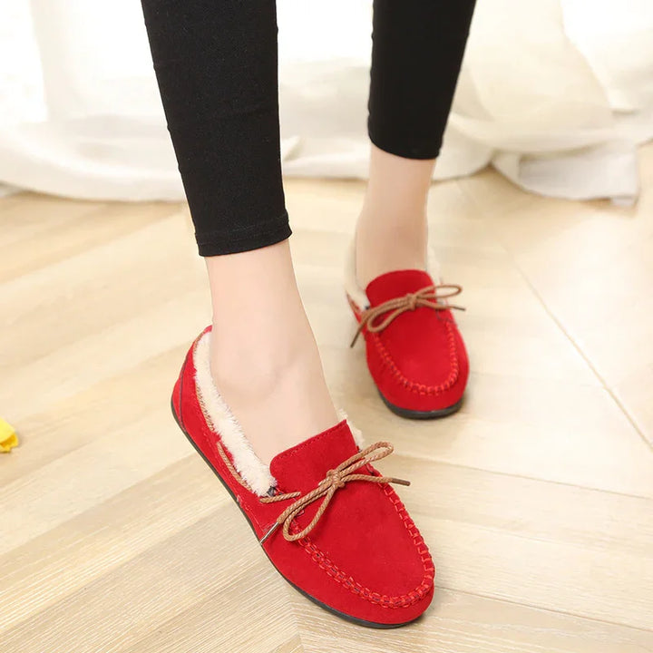Red suede moccasin slippers with fur lining and brown bow detail worn with black pants on wooden floor