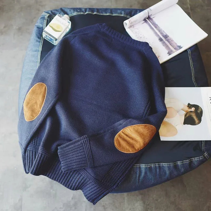 Navy blue knitted sweater with brown elbow patches on a navy cushion, surrounded by a magazine and perfume bottle