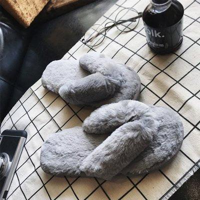 Pair of soft gray plush slippers on a checked cloth with glasses, black bottle, and phone nearby
