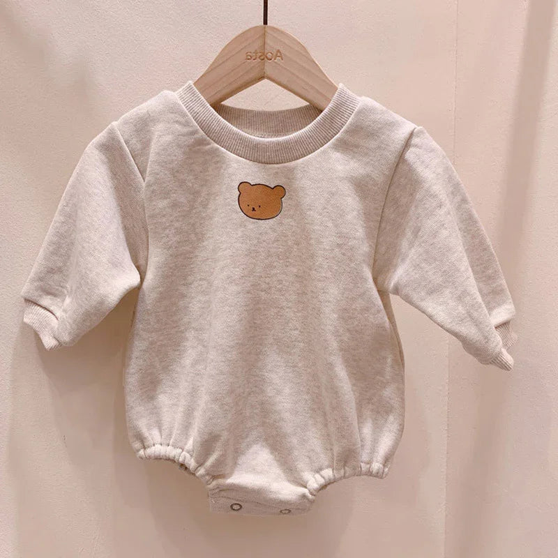 Beige baby romper with long sleeves and cute bear face print on front, hanging on wooden hanger
