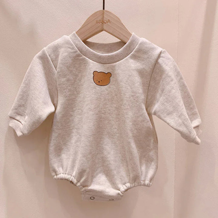 Beige baby romper with long sleeves and cute bear face print on front, hanging on wooden hanger