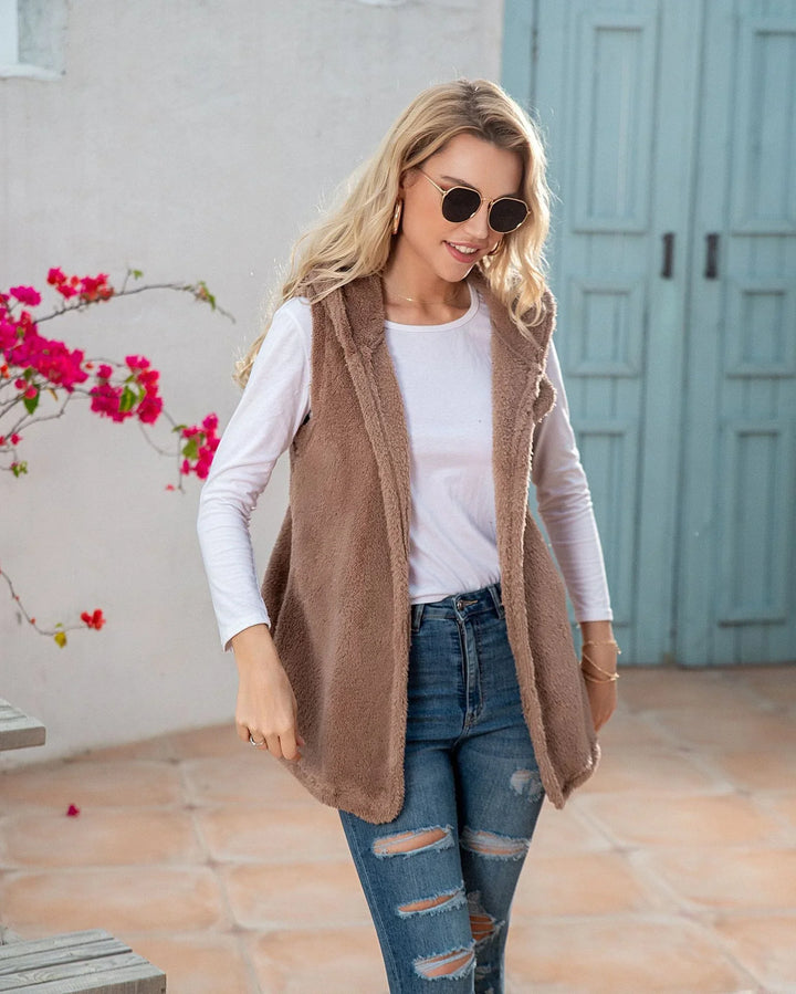 Blonde woman wearing sunglasses, white long-sleeve shirt, brown fuzzy vest, and ripped jeans in outdoor courtyard