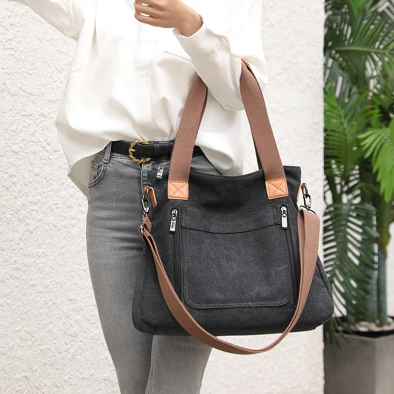 Black canvas tote bag with brown handles and adjustable strap worn by person in white top and gray jeans