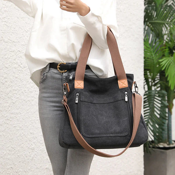 Black canvas tote bag with brown handles and adjustable strap worn by person in white top and gray jeans