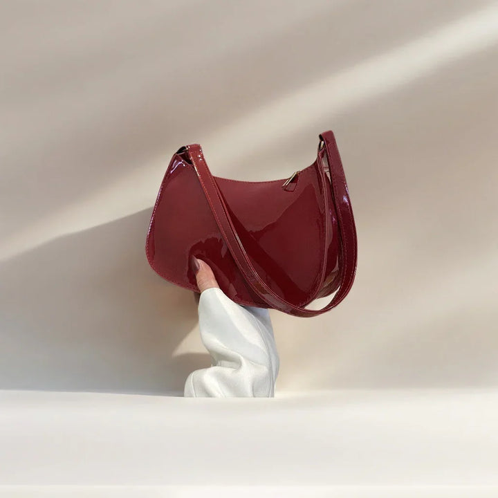 Hand holding a shiny dark red patent leather shoulder bag against a beige background